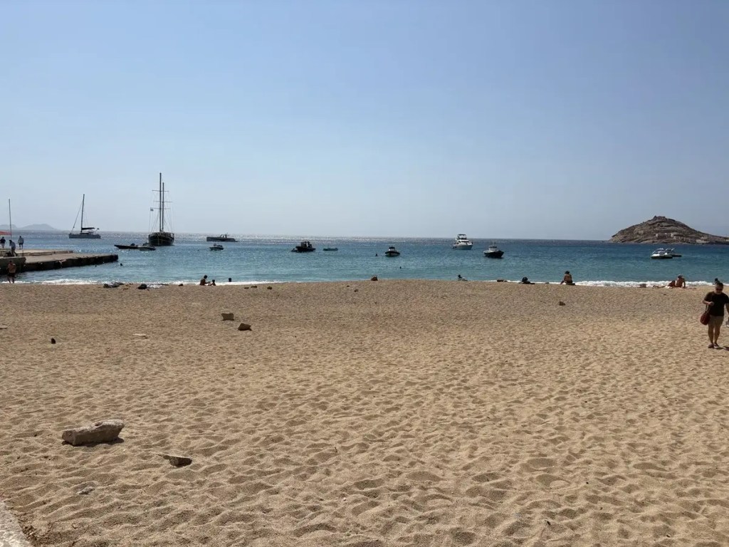 Sandy beach facing turquoise Aegean water with sailboats and small boats near Mykonos