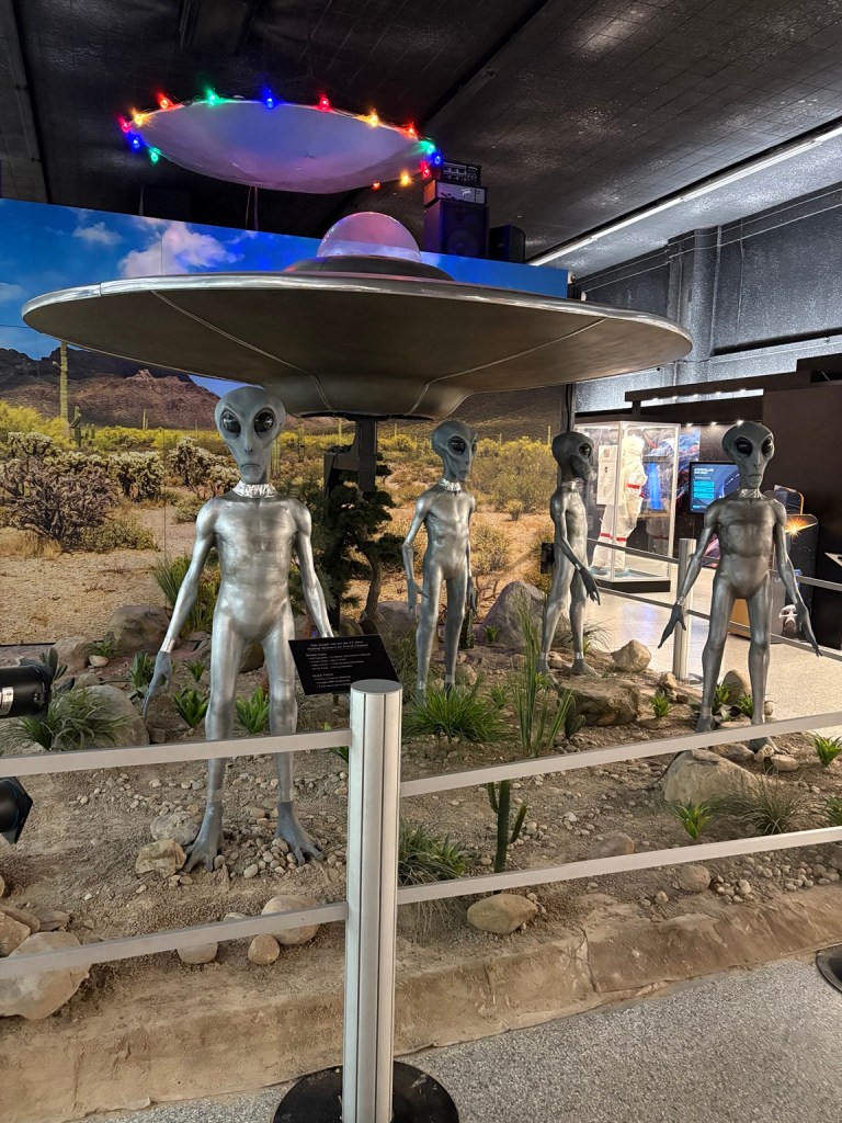 Four alien figures standing beneath a flying saucer exhibit at the International UFO Museum.