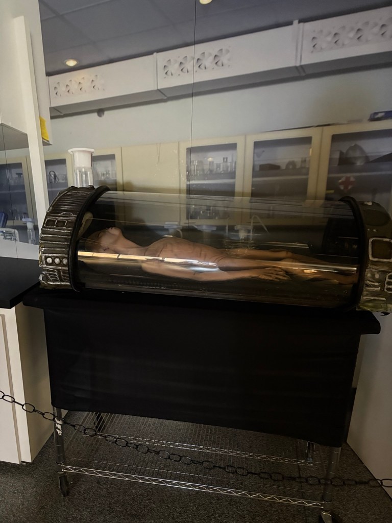 Alien figure lying inside a transparent science-fiction-style capsule display.