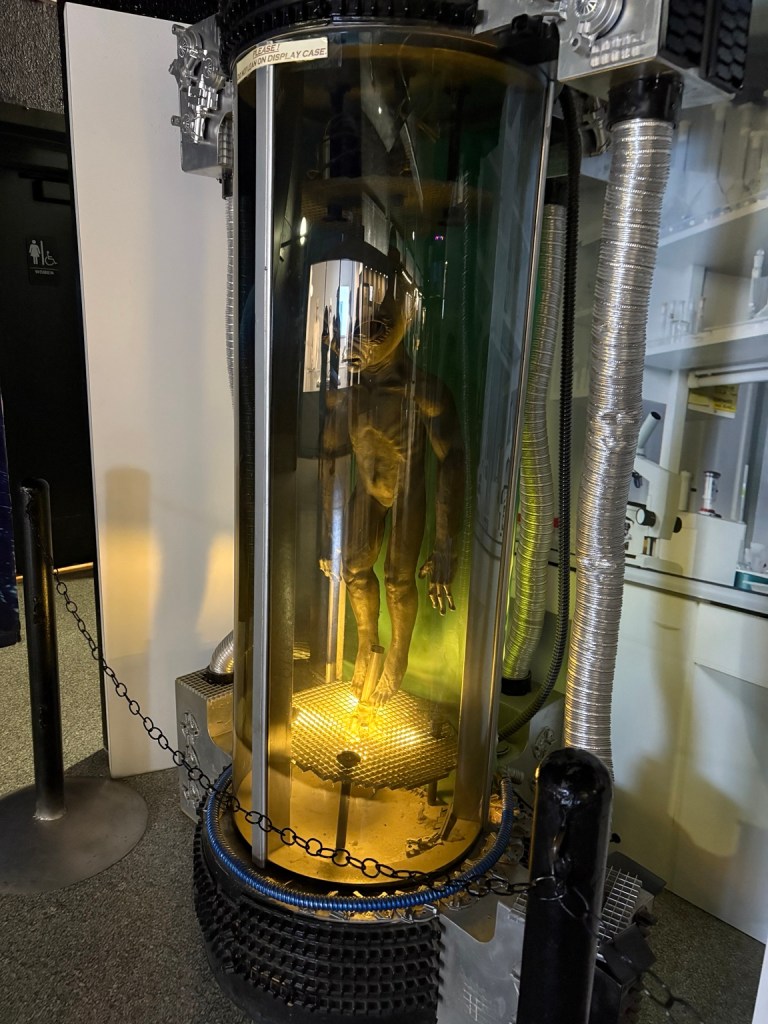 Alien figure inside a glowing cylindrical science-fiction-style display.
