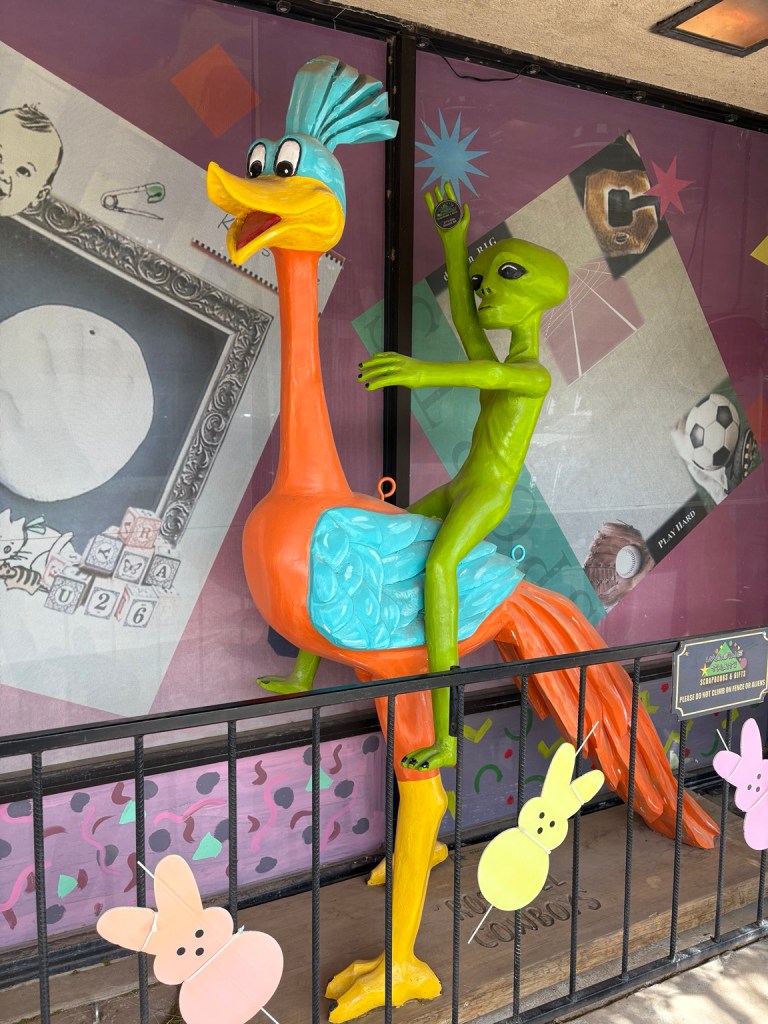 Bright green alien riding a colorful oversized bird outside a Roswell shop.