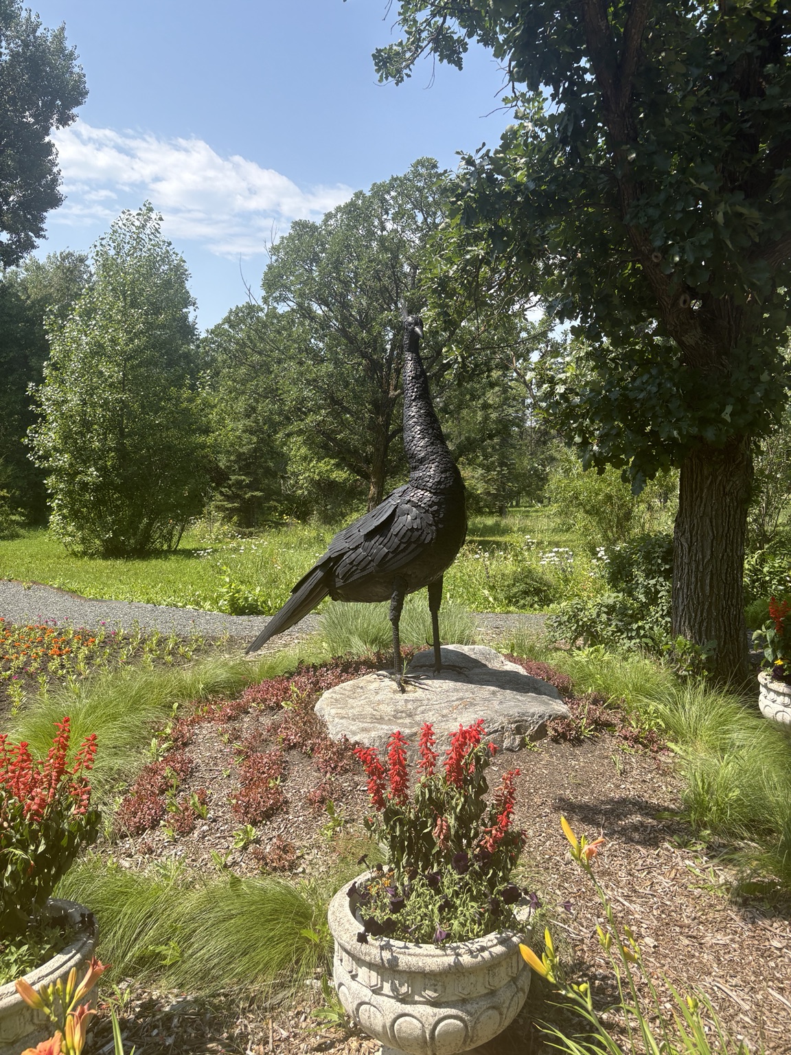 A dark metal bird sculpture stands on a rock among flowers and trees.