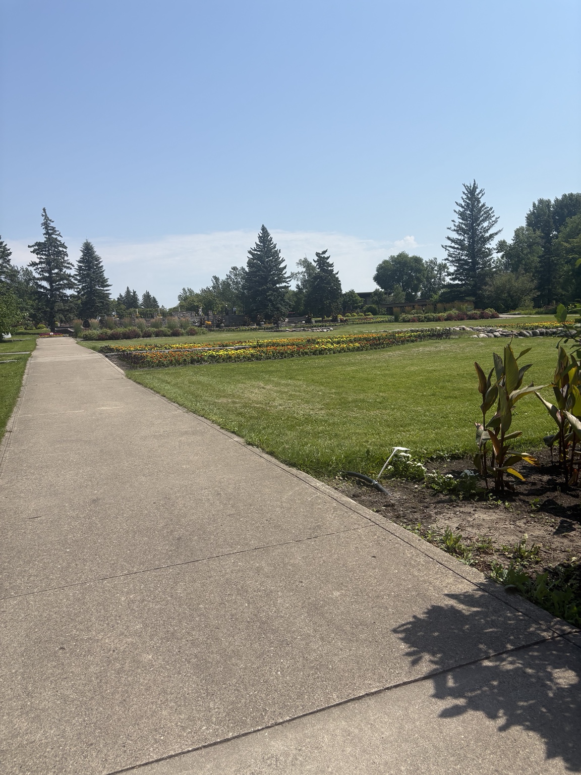 A paved walkway beside lawns, colorful flower beds, and trees at the International Peace Garden.