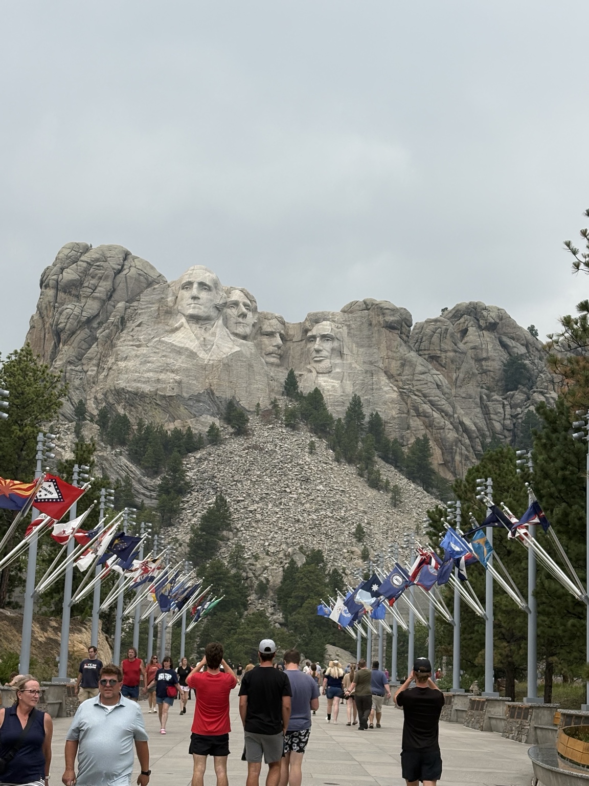 Visitors walk between rows of state flags toward Mount Rushmore beneath an overcast sky.